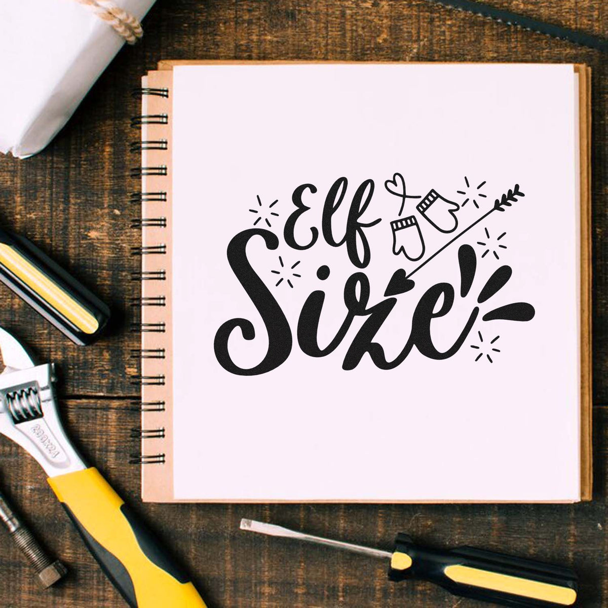 Black Elf Size! stamp and seal design on a white spiral notebook, with mitten and arrow graphics, beside craft tools.