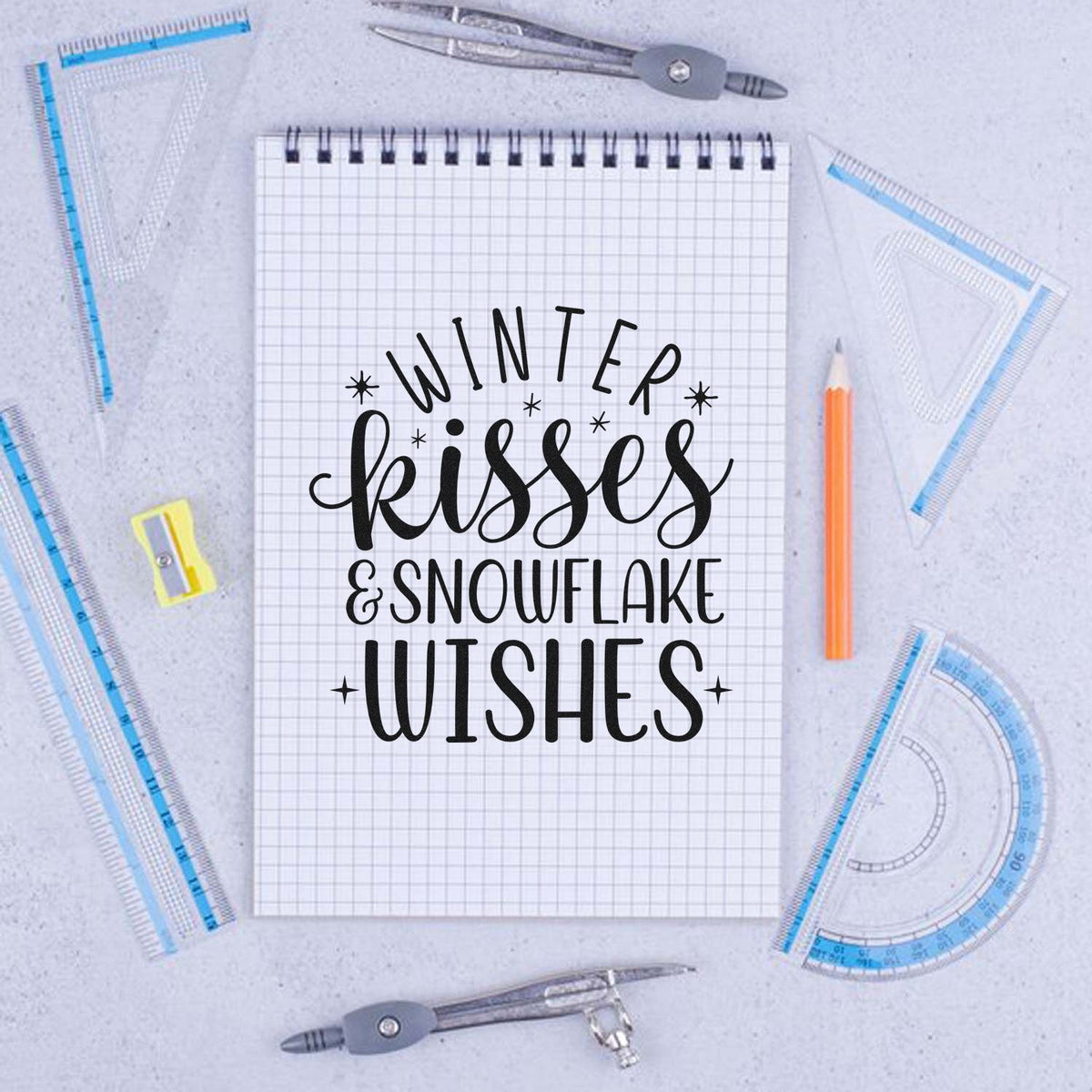 Black Winter Kisses & Snowflake Wishes stamp on graph notebook, framed by blue drafting tools and an orange pencil.