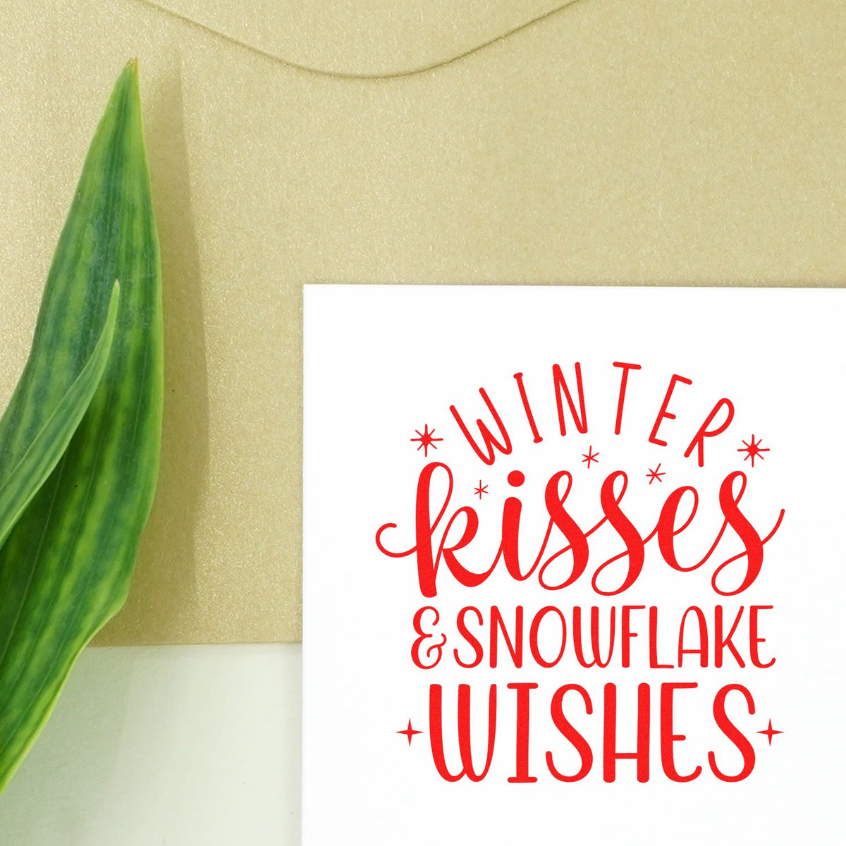 Red Winter Kisses & Snowflake Wishes stamp on white card, beside a green leaf on beige background.