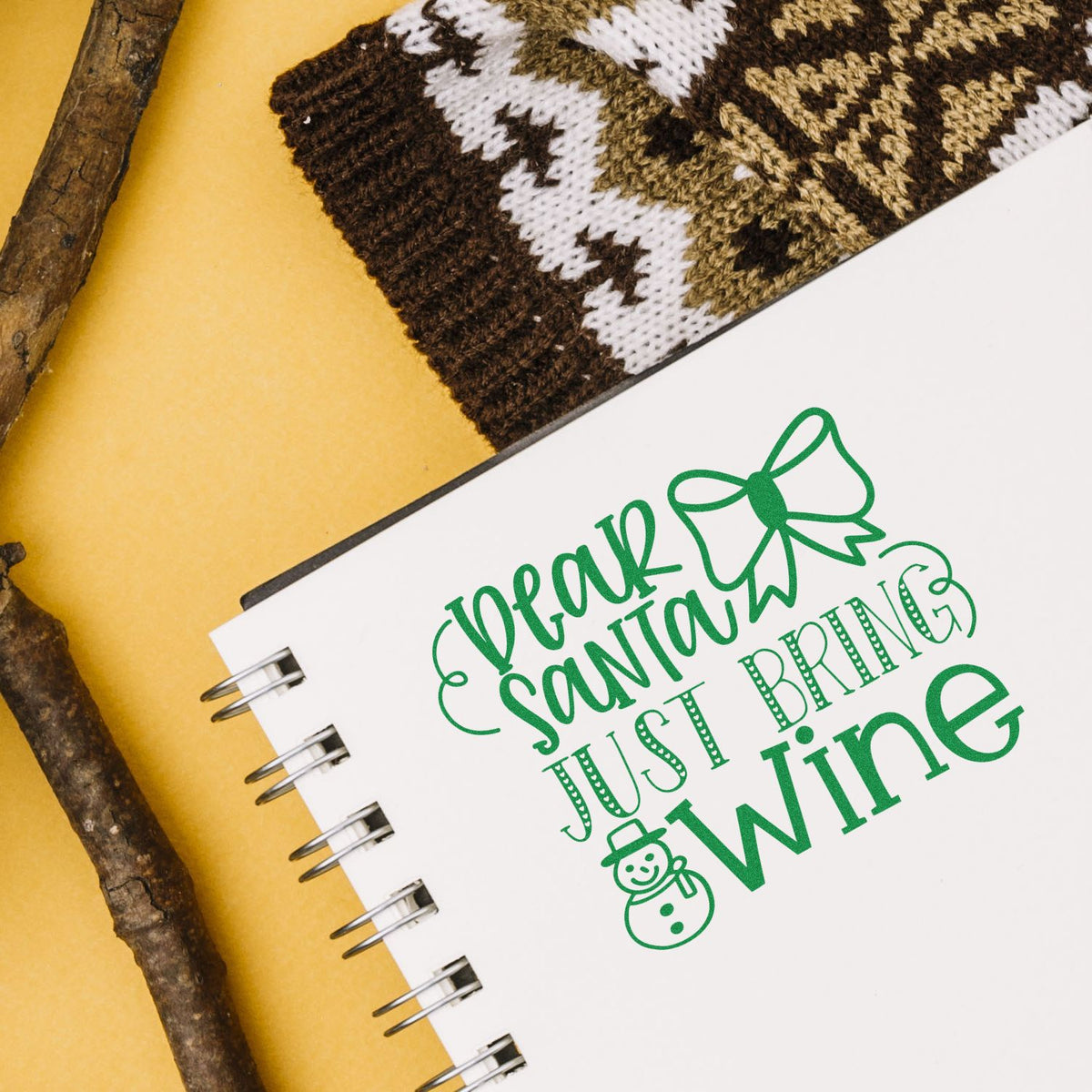 Green holiday stamp and seal reading Dear Santa Just Bring Wine on white notebook, with knitted stocking and rustic twigs.