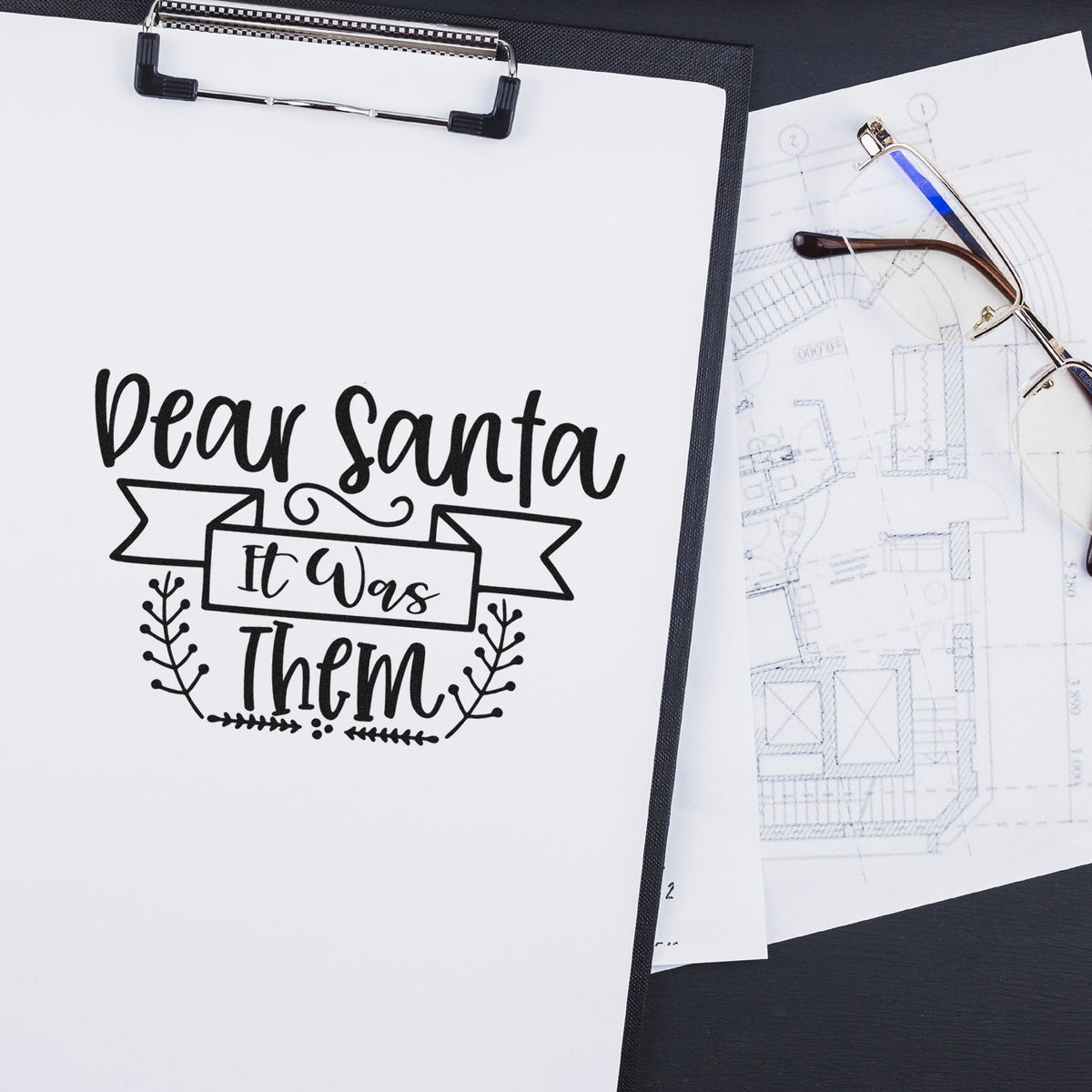 Black custom stamp and seal reading Dear Santa It Was Them on white paper beside clipboards, blueprint and eyeglasses.