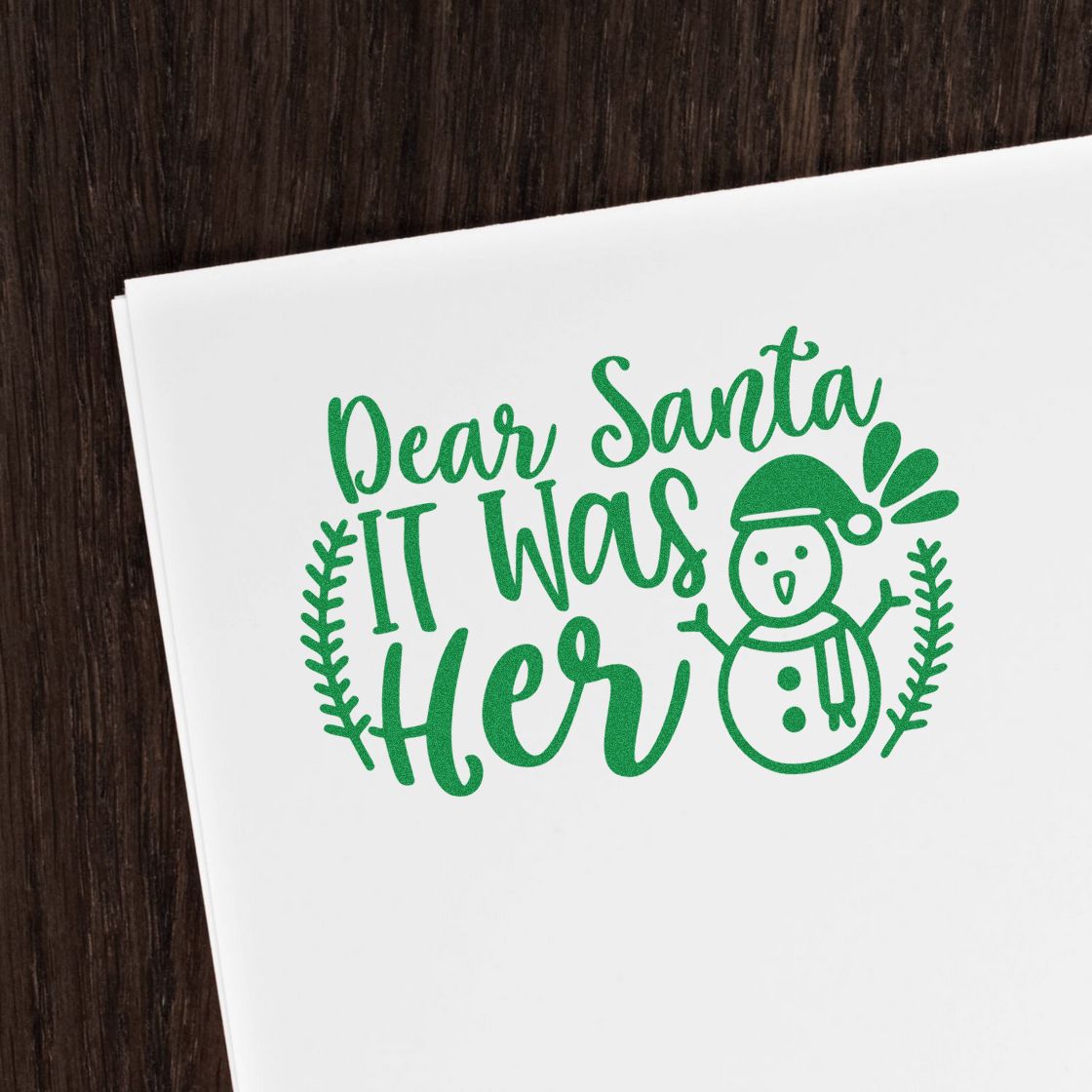 Green holiday stamp and seal design on white paper, reading Dear Santa It Was Her with a snowman and wreath.