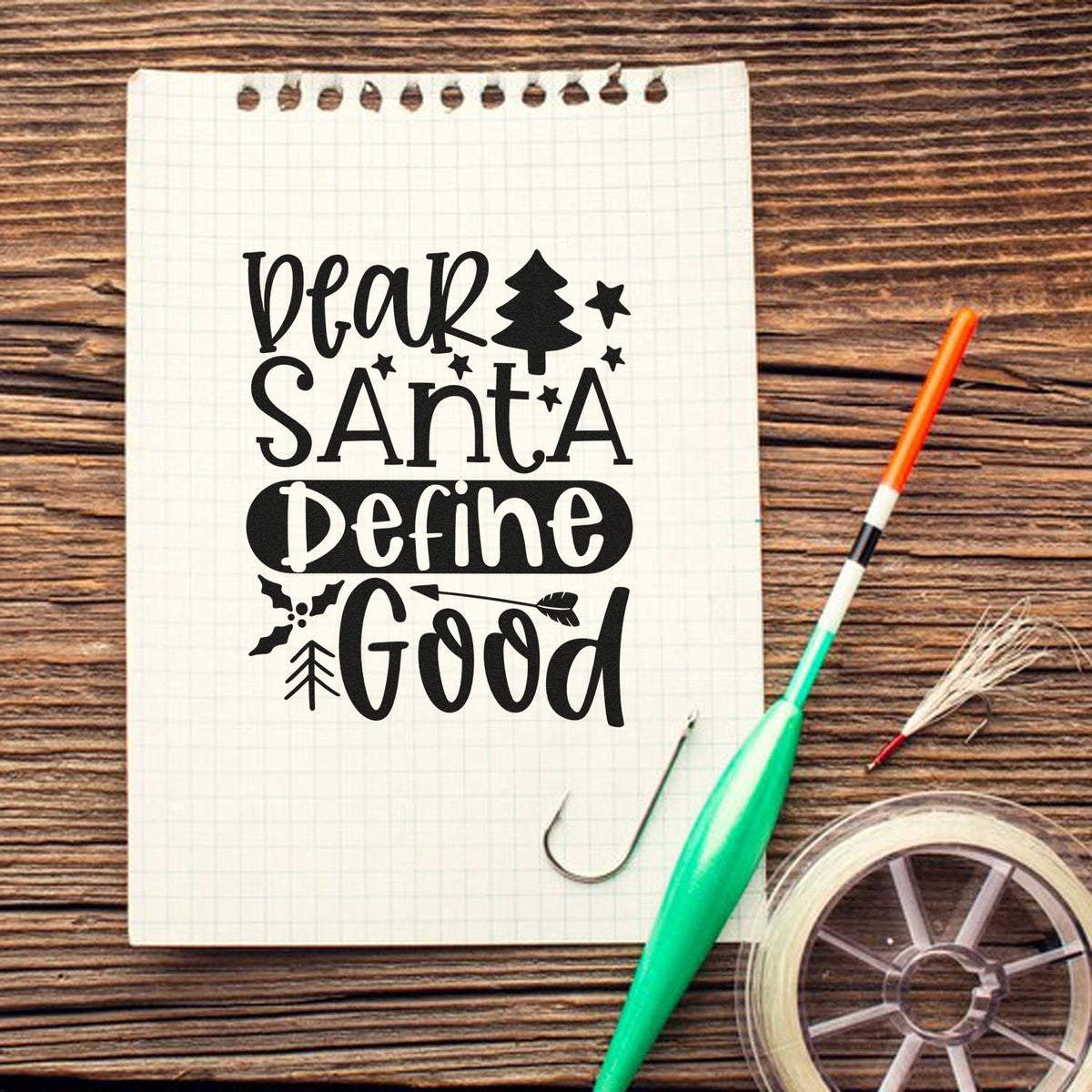 Black Christmas stamp and seal reading Dear Santa Define Good with tree and stars on a white grid notebook, wood table.