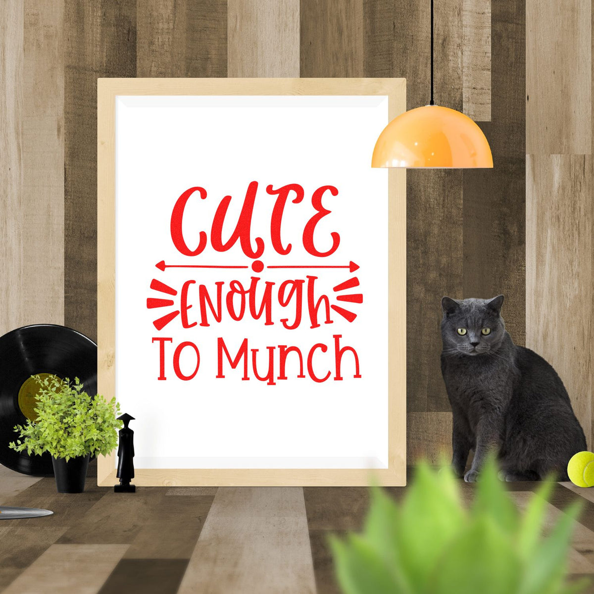 Framed stamp/seal-style print with red Cute Enough lettering on white, styled on a wood wall beside a black cat.