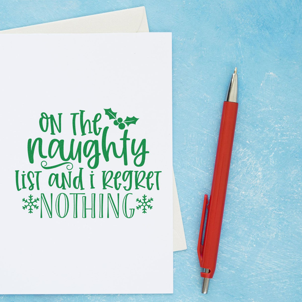 Green holiday stamp and seal on white card reads On the naughty list and I regret nothing, with holly, snowflakes, red pen, blue background.