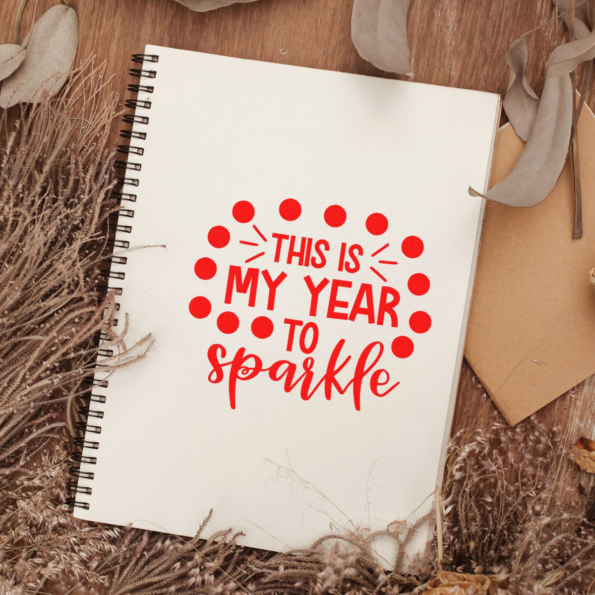 Red stamp and seal reading This is my year to sparkle on a cream spiral notebook with dried beige florals.