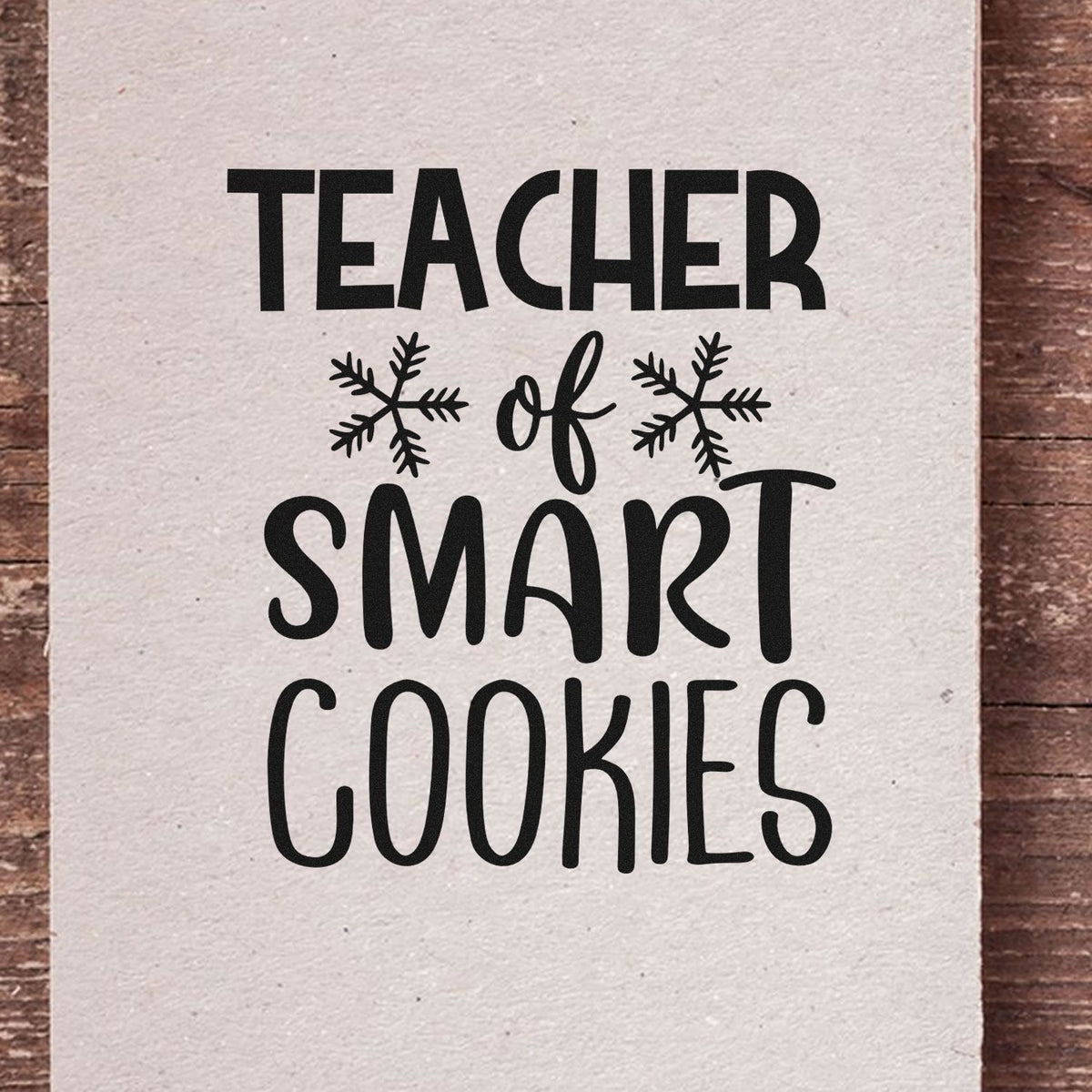 Black teacher stamp design reading Teacher of Smart Cookies with snowflake accents on cream paper and wood background.