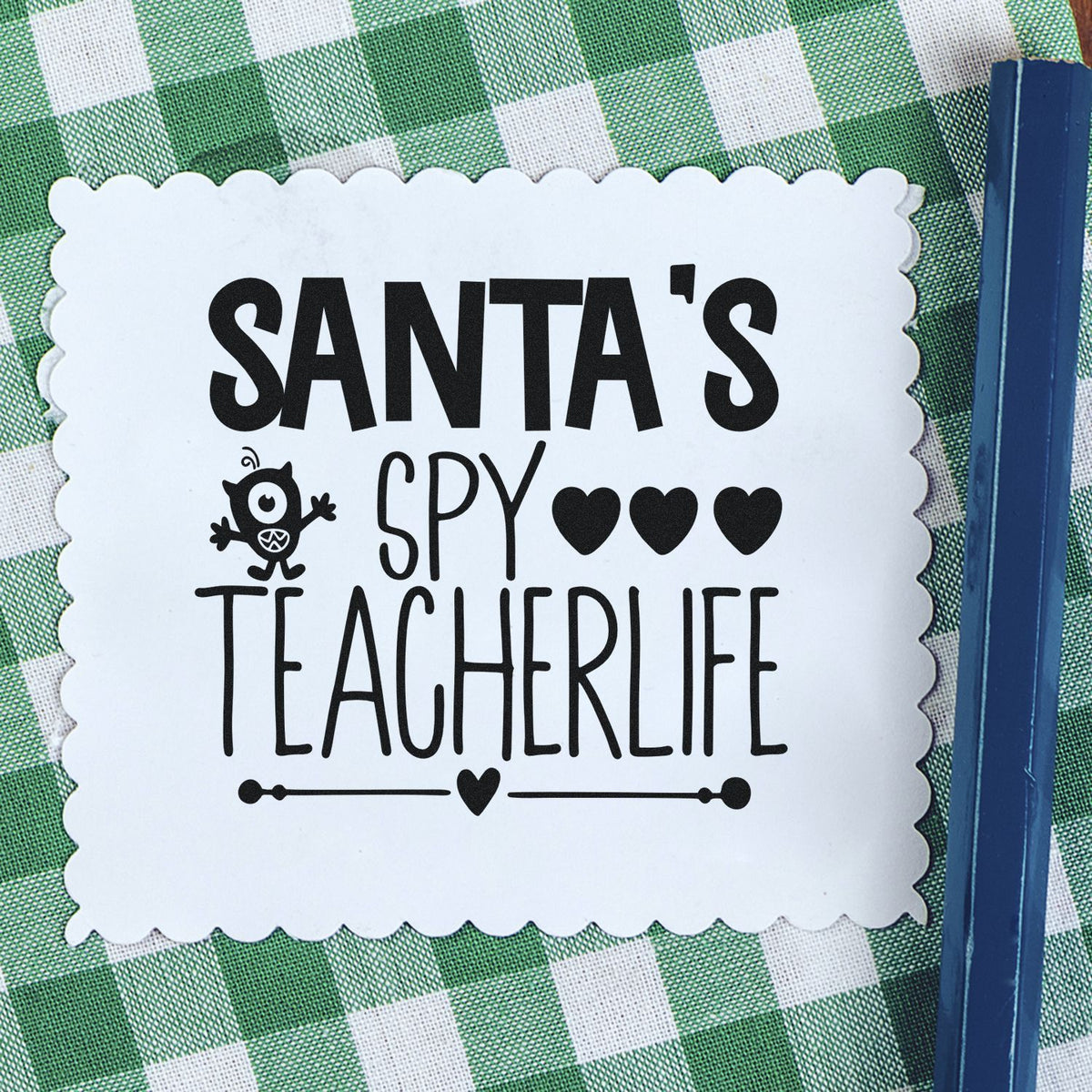 White scalloped stamp seal with black Santa's Spy Teacher Life text, hearts, and monster icon on green gingham cloth.