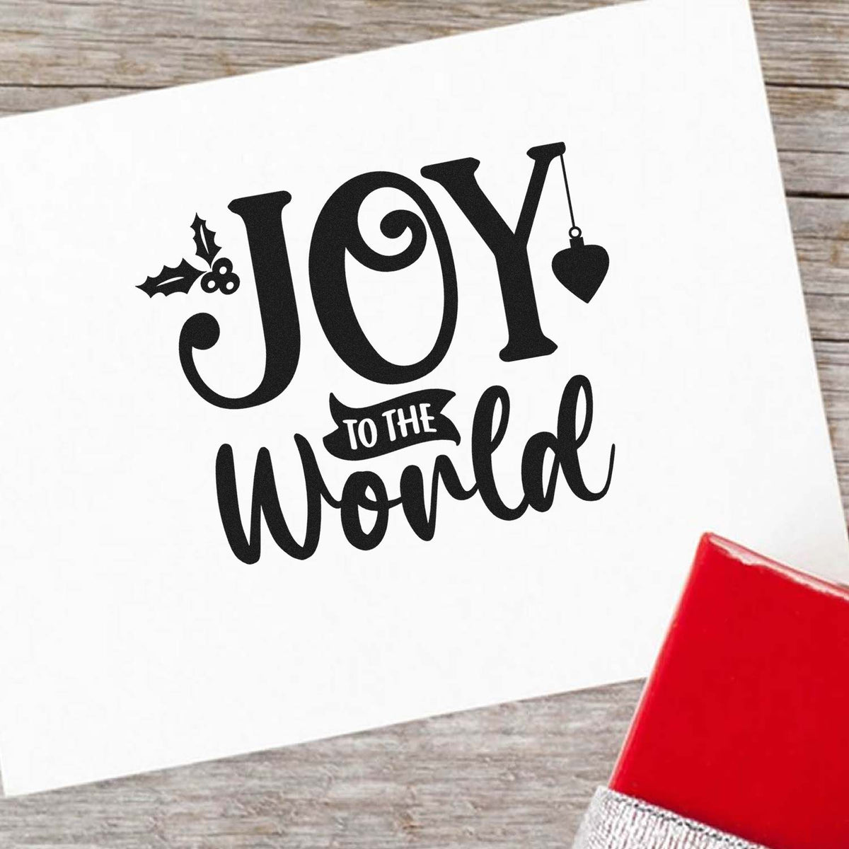 Black Joy to the World holiday stamp and seal design with holly, heart ornament, on white paper beside a red gift box.