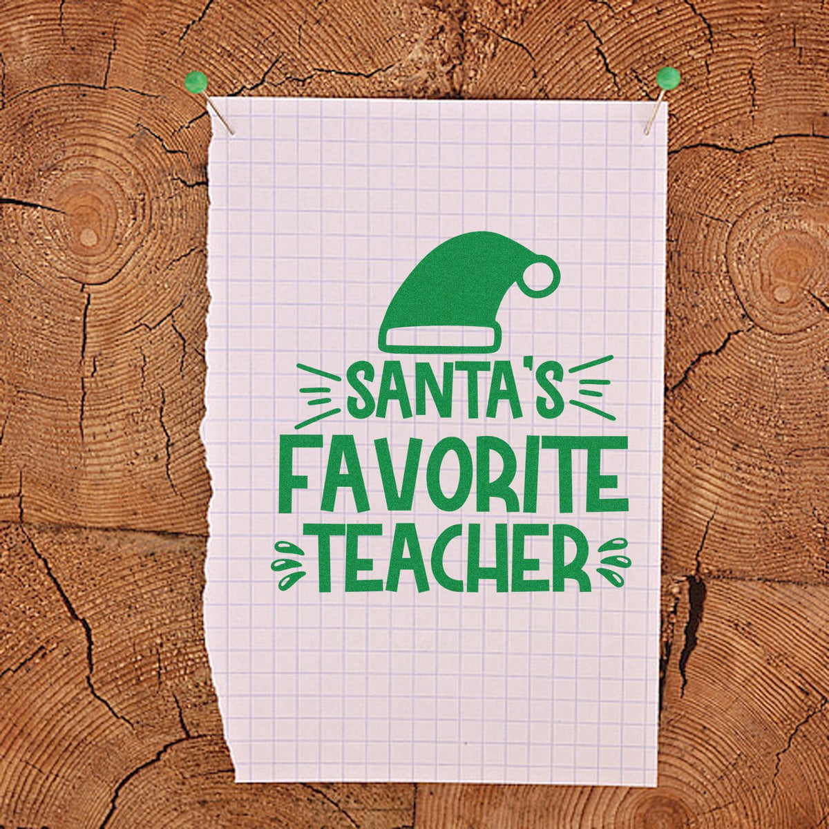 Green Santa hat stamp and seal reading Santas Favorite Teacher on white grid paper with wood background.