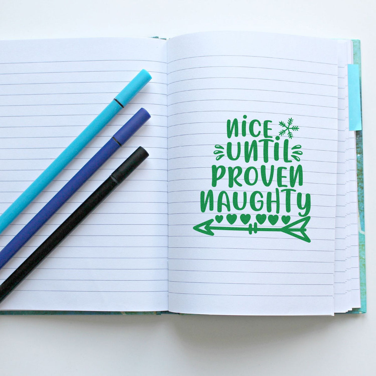 Open lined notebook with a green holiday stamp-seal reading Nice Until Proven Naughty beside blue, purple, and black pens.