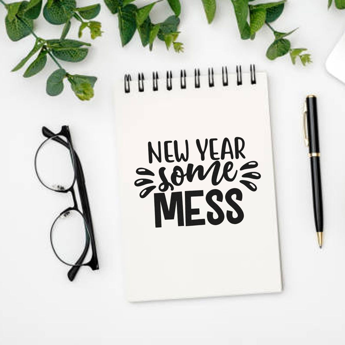 New Year Some Mess stamp design on white spiral notebook, with black pen, glasses, and green leaves.