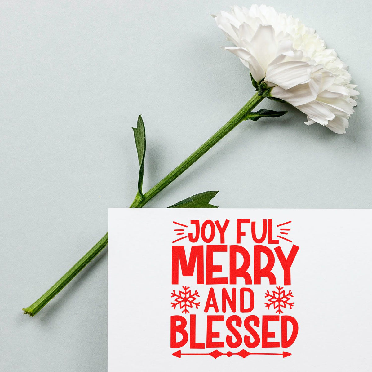 White flower beside a red holiday stamp and seal reading Joyful Merry and Blessed on white paper.