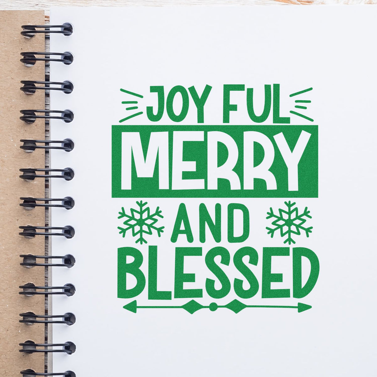 Green holiday stamp and seal design reading Joyful, Merry and Blessed with snowflakes on a white spiral notebook.