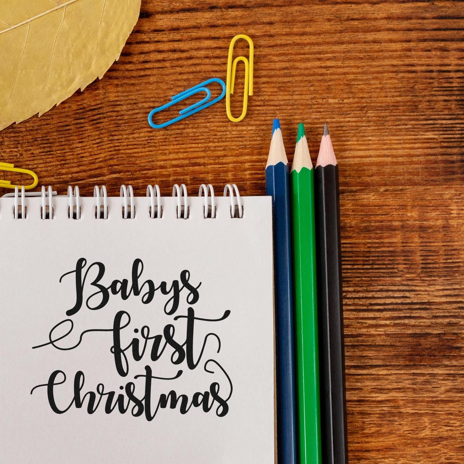 Babys first Christmas stamp and seal design on white spiral notebook with blue, green, black pencils and colorful paper clips.