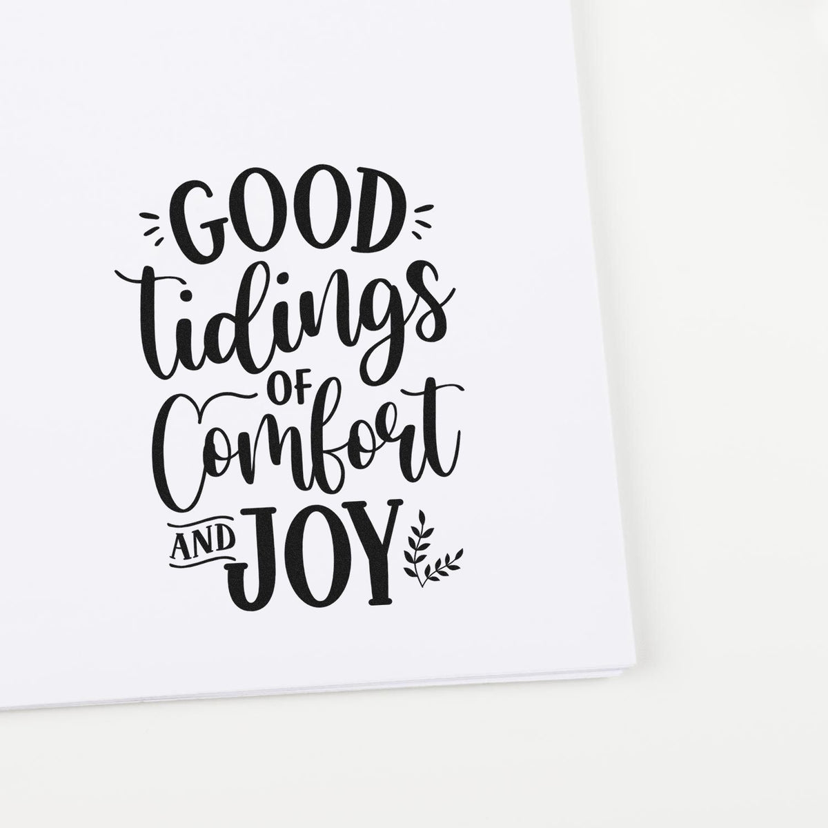 Black decorative stamp reading Good Tidings of Comfort and Joy on white paper with leaf accents.