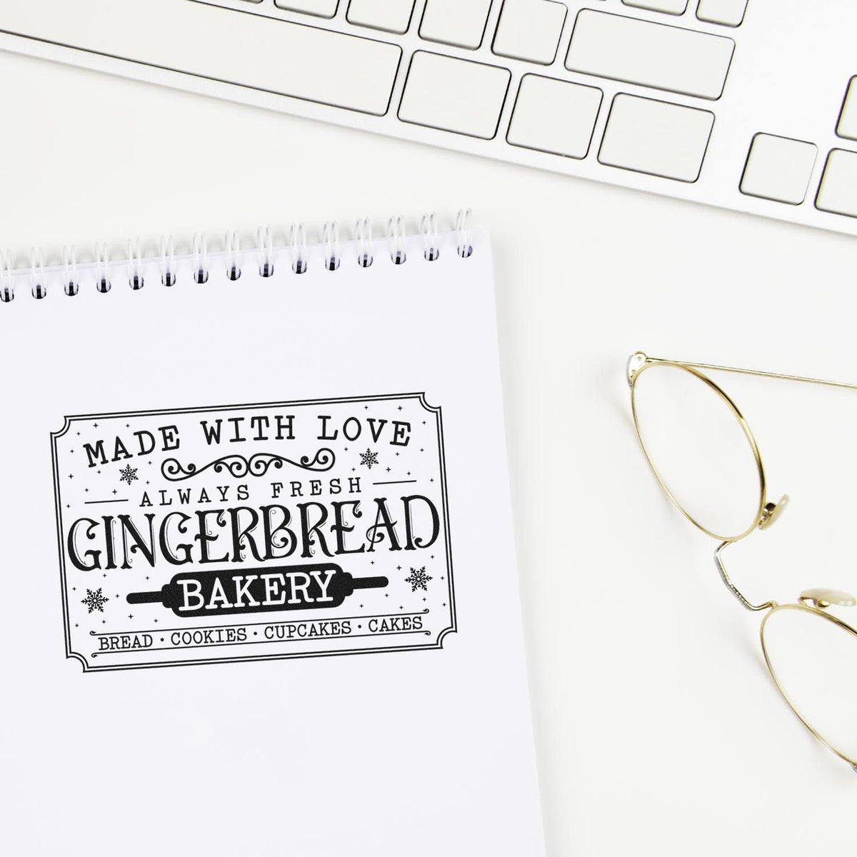 Black-and-white gingerbread bakery stamp seal on a white spiral notebook, beside a white keyboard and gold glasses.
