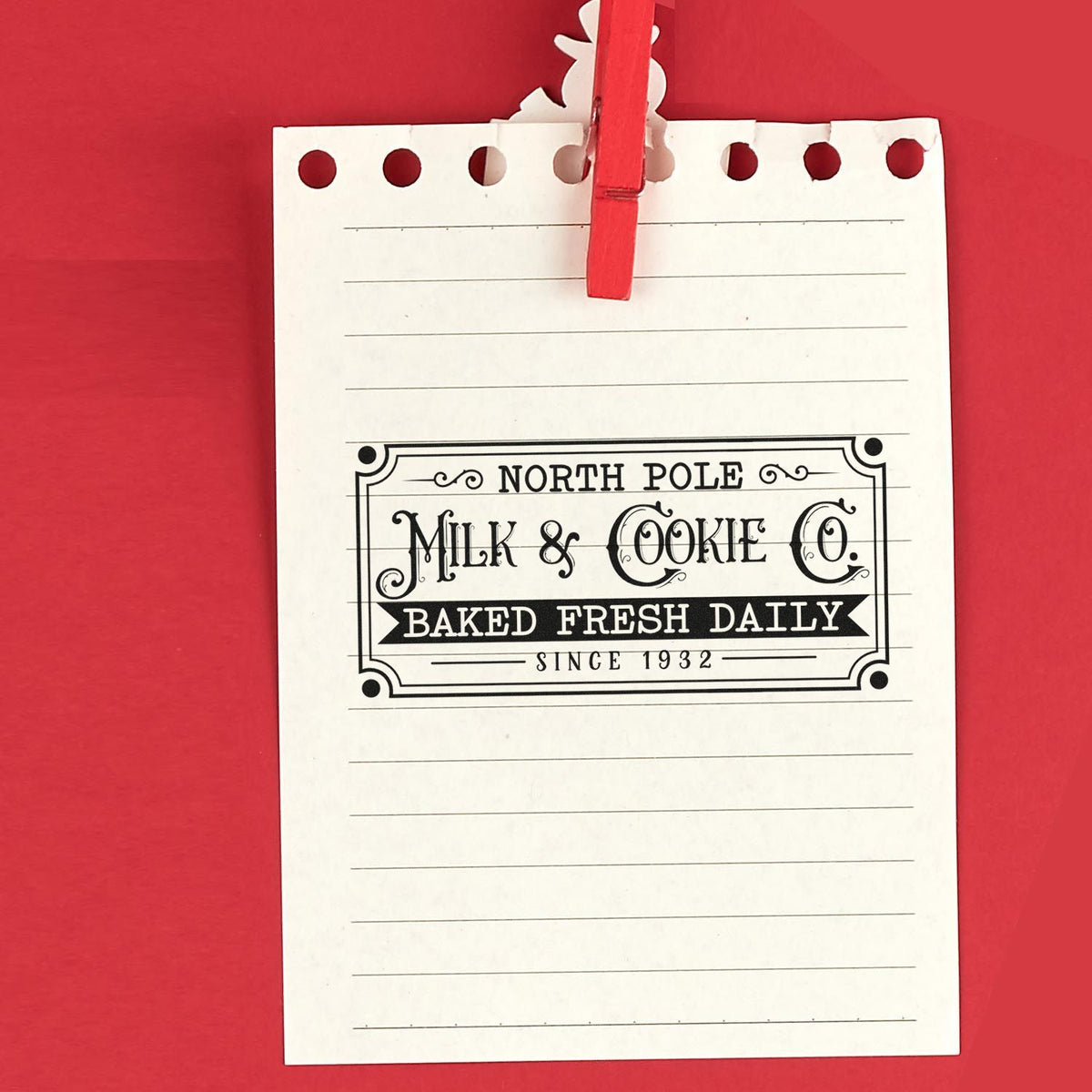 White notepad with red clothespin on red background, featuring a vintage black North Pole Milk & Cookie Co. stamp seal.