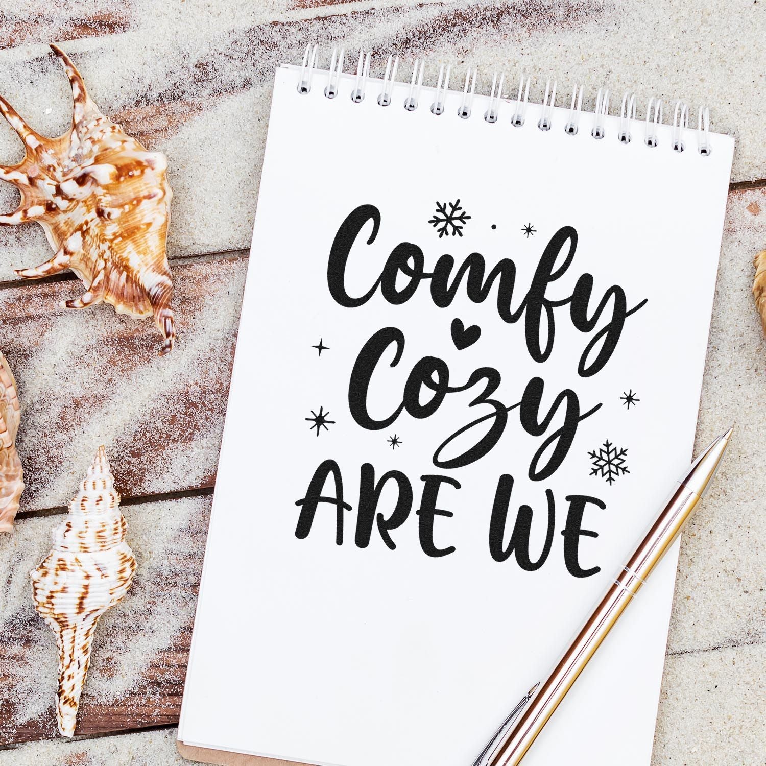 Black custom stamp and seal reading Comfy Cozy ARE WE on a white spiral notebook, with snowflakes, seashells and gold pen.