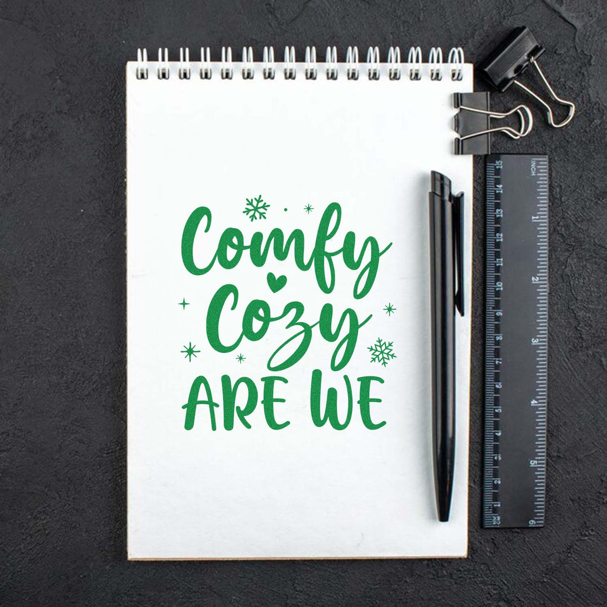 Green Comfy Cozy Are We stamp design with snowflakes and heart on white spiral notebook, beside black pen, ruler, and binder clip.