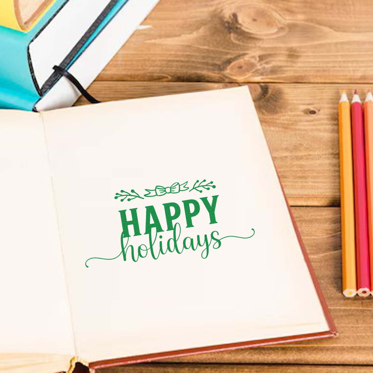 Green Happy Holidays stamp and seal on open notebook, with colorful pencils on a wooden desk.
