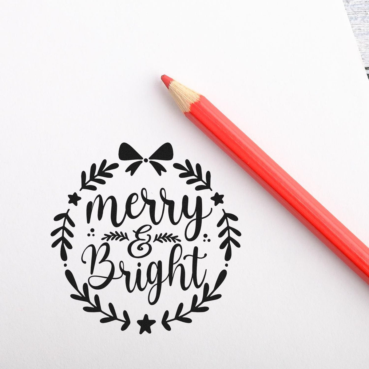 Black Merry & Bright wreath stamp seal beside a red pencil on white paper.