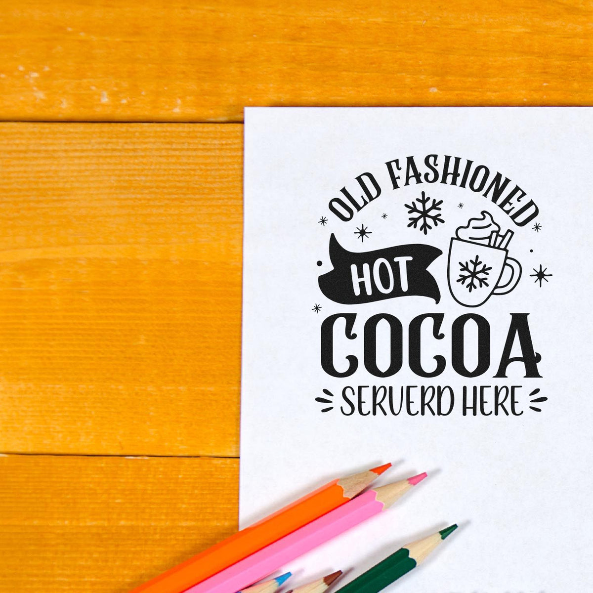 Black hot cocoa stamp and seal on white paper, with mug and snowflakes, on orange wood and colorful pencils.