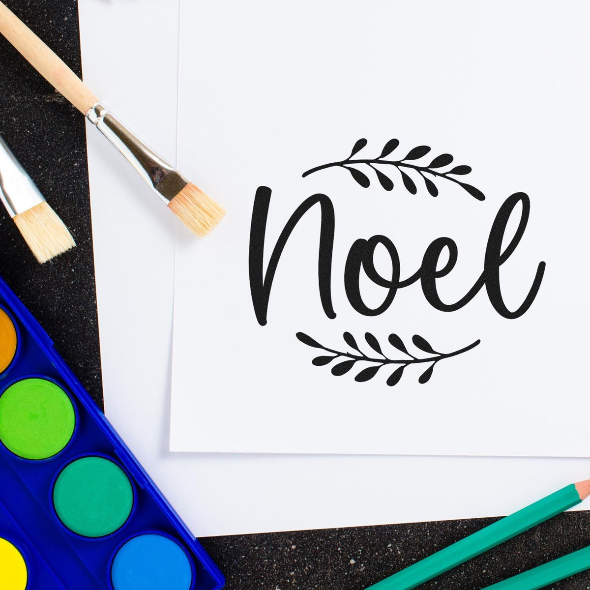 Black Noel stamp and seal product on white card, with paintbrushes, watercolor palette, and teal pencils.