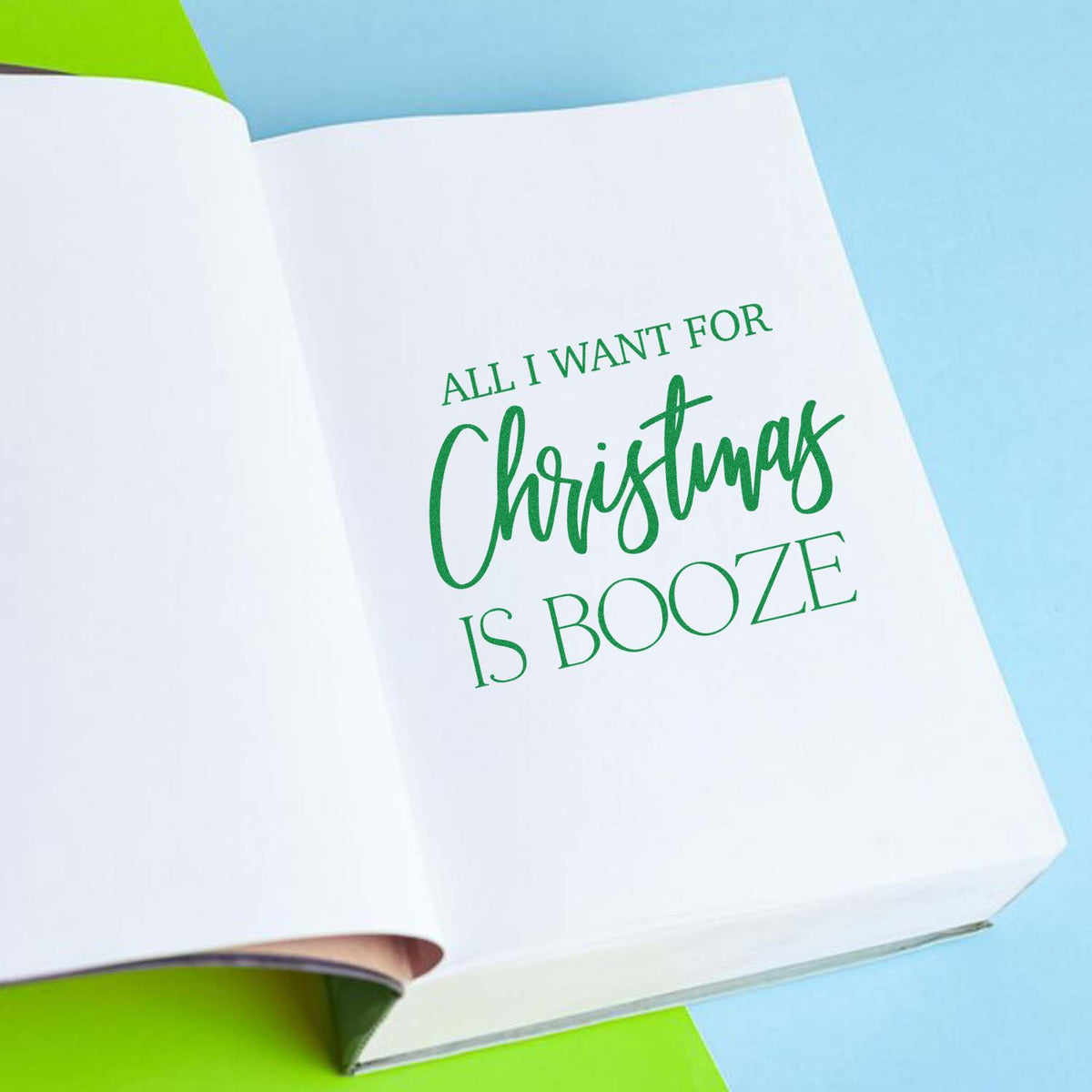 Green Christmas stamp and seal design reading All I want for Christmas is booze on white paper over a blue background.