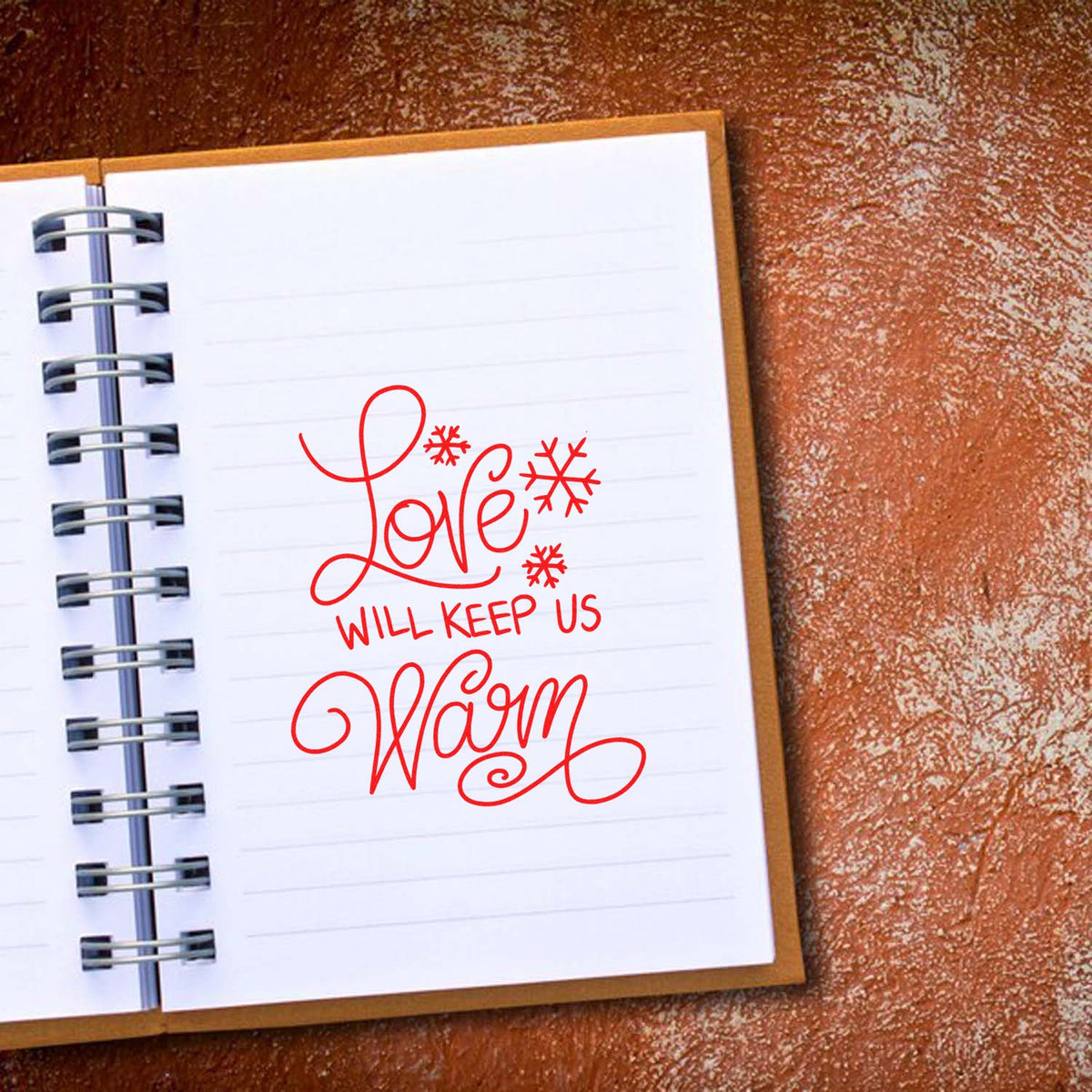 Red decorative stamp reading Love will keep us warm with snowflakes on a white spiral notebook, on a brown textured background.