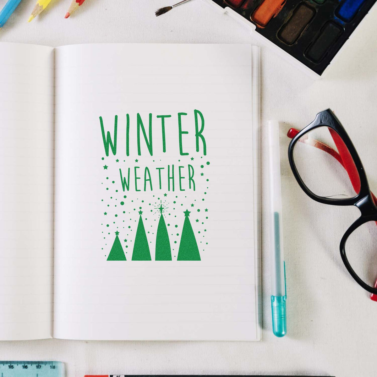 Green Winter Weather stamp/seal design on open notebook, with Christmas tree silhouettes, stars, and art supplies nearby.