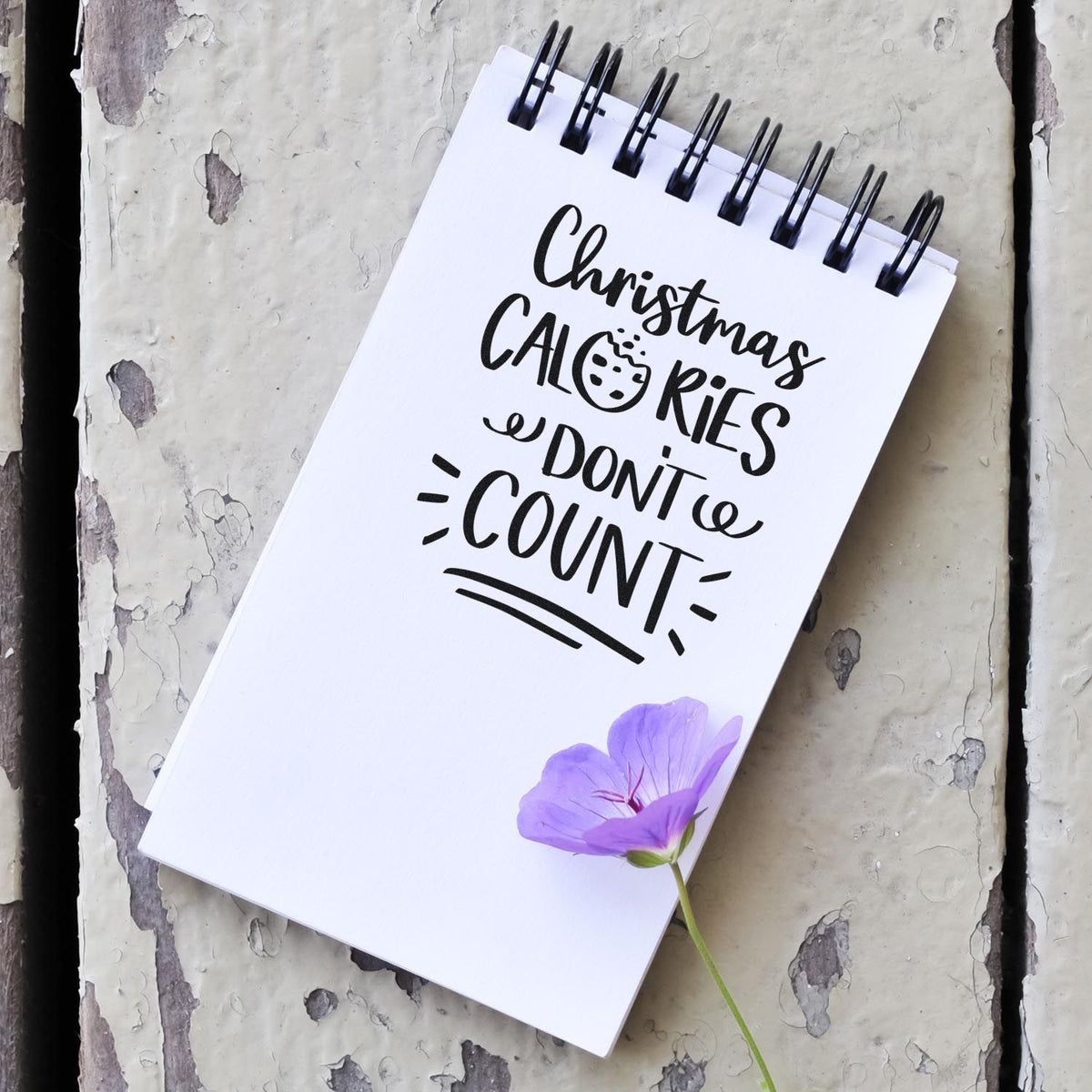 White spiral notebook with black Christmas calories don't count stamp/seal, purple flower, on distressed gray surface.