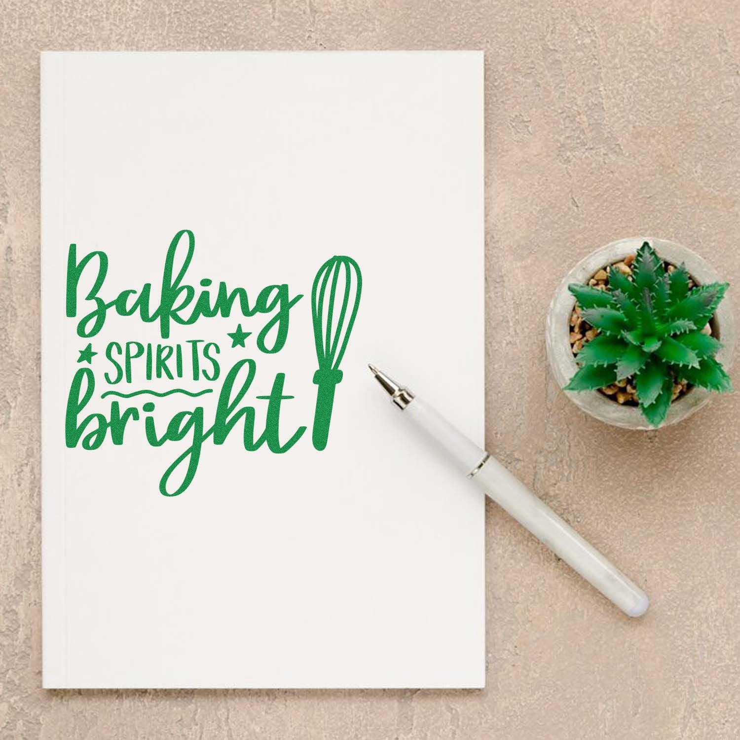 Green Baking Spirits Bright stamp and seal design on white notebook with whisk graphic, white pen, and small succulent.