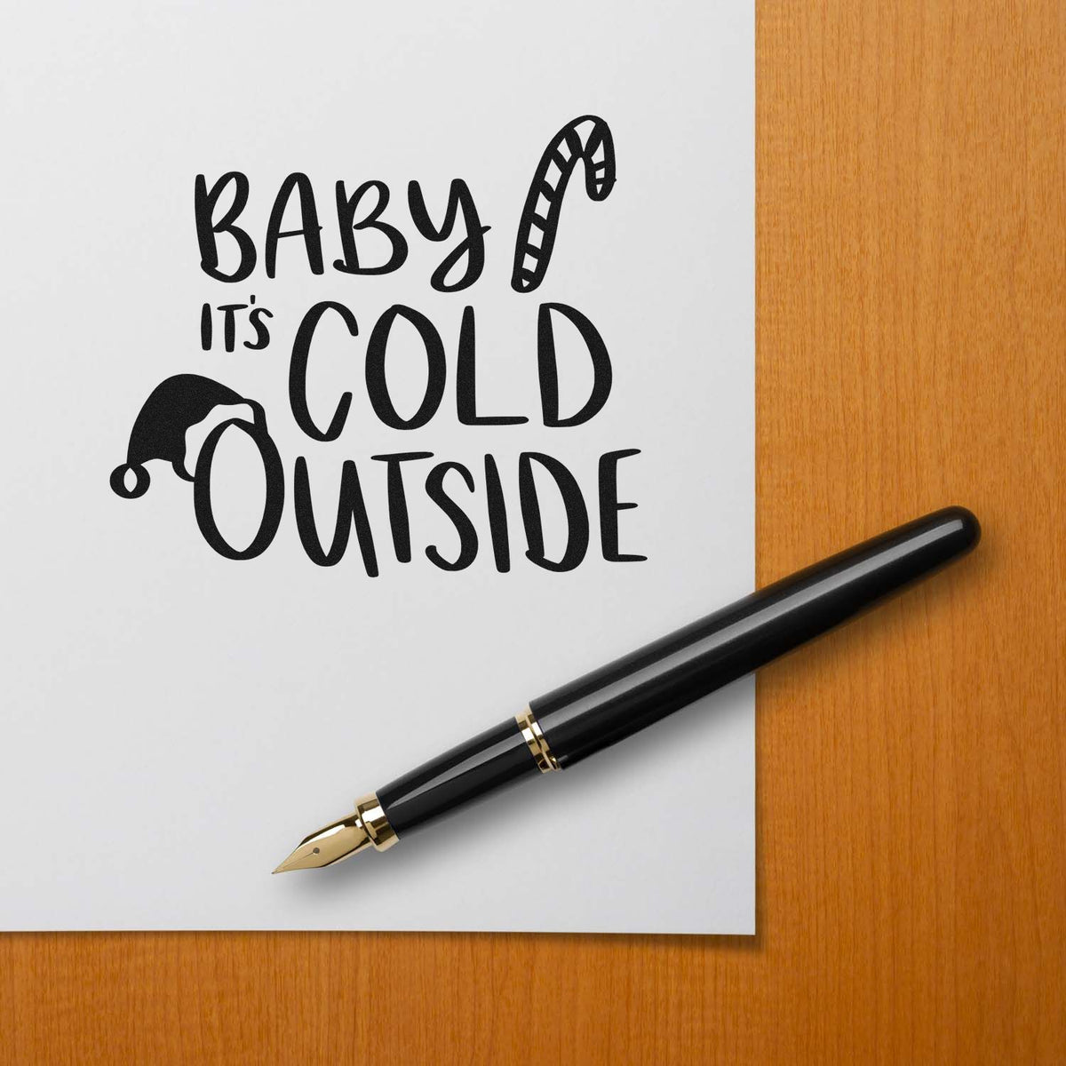 Black holiday stamp/seal design reading Baby its cold outside with candy cane and Santa hat, beside a black fountain pen.