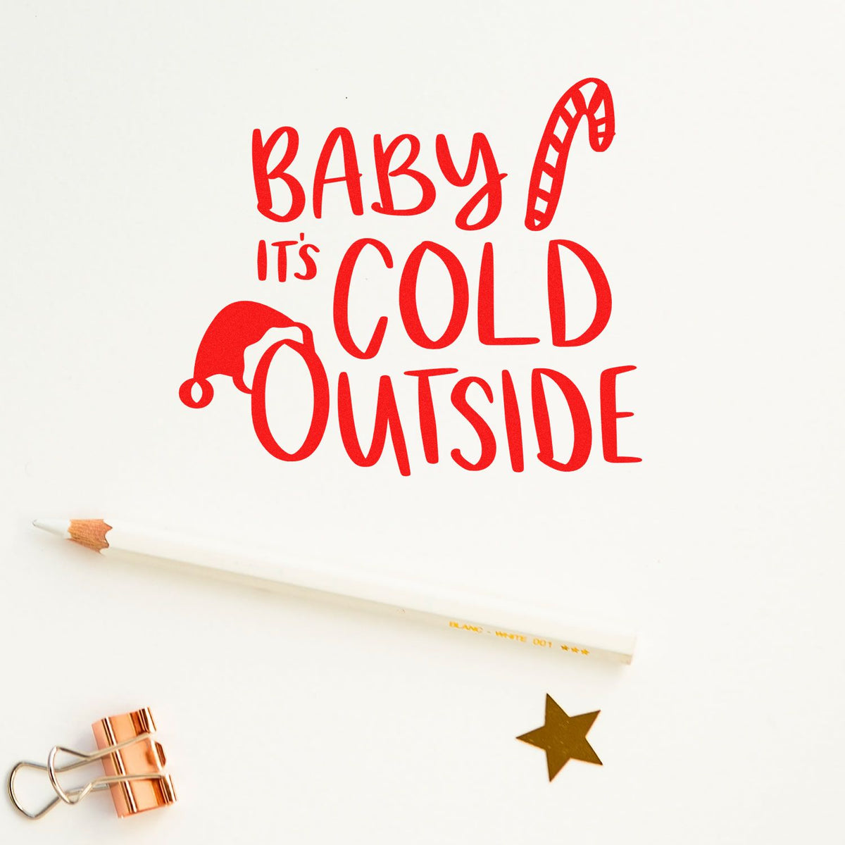 Red holiday stamp and seal reading 'Baby Its Cold Outside' with candy cane, Santa hat, white pencil, gold star, and rose gold clip.