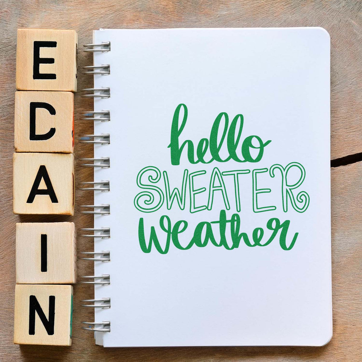 Wooden alphabet stamp blocks beside a white spiral notebook stamped hello SWEATER weather in green, showcasing stamp and seal products.