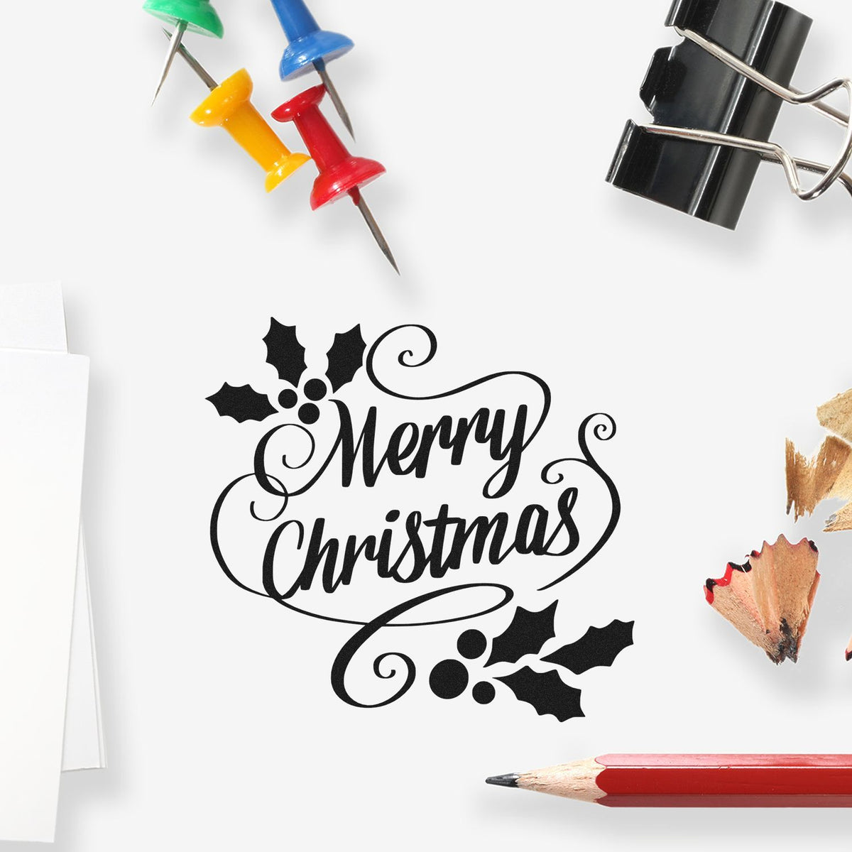 Black Merry Christmas decorative stamp and seal with holly accents, surrounded by colorful push pins, pencil, and binder clip.