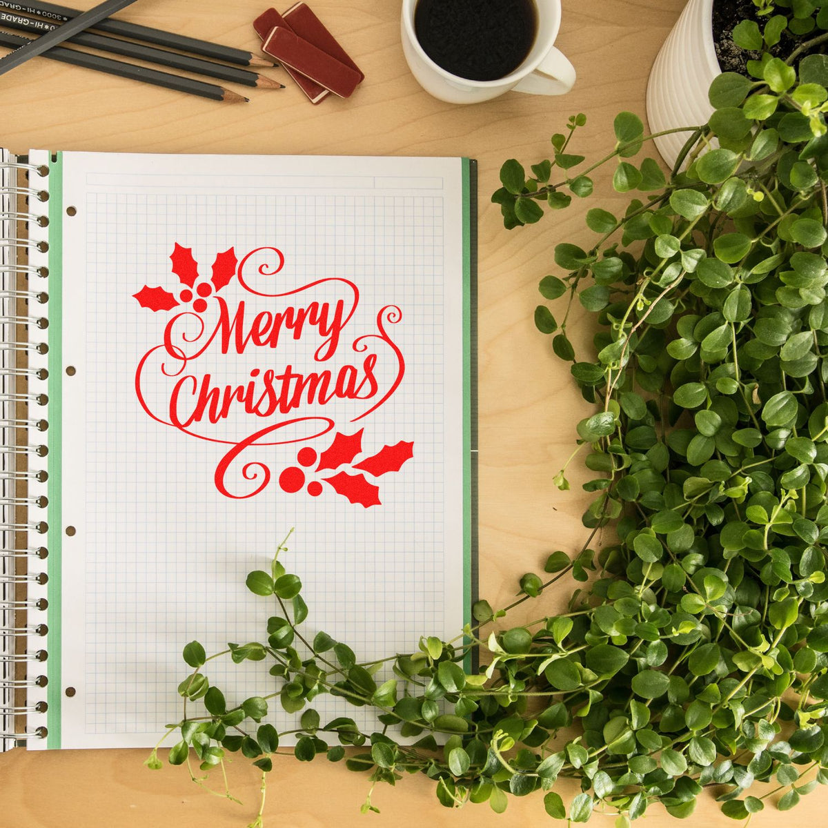 Red Merry Christmas stamp and seal with holly on graph paper notebook, beside green plant, coffee cup, and pencils.