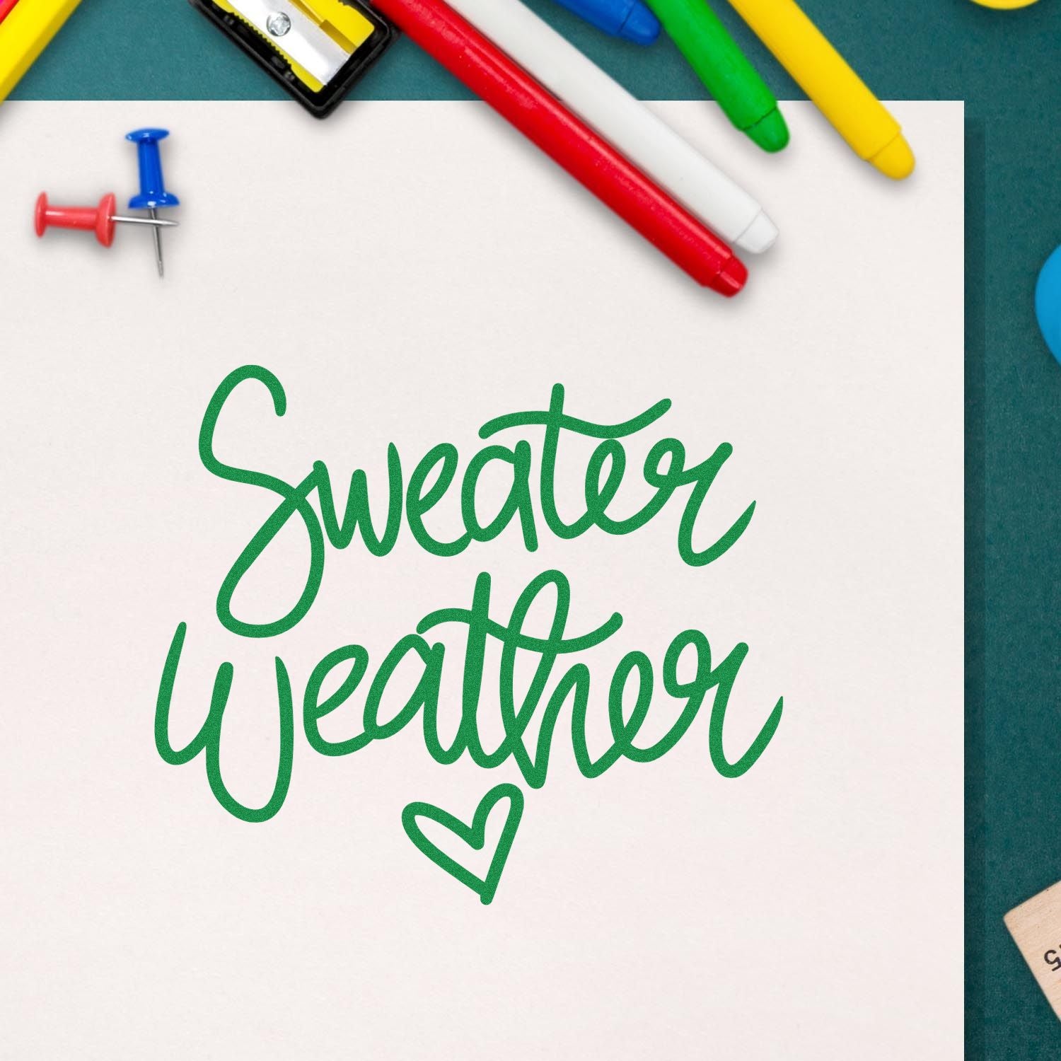 Green stamp-and-seal reading Sweater Weather on white paper, with a heart and colorful markers on a teal background.