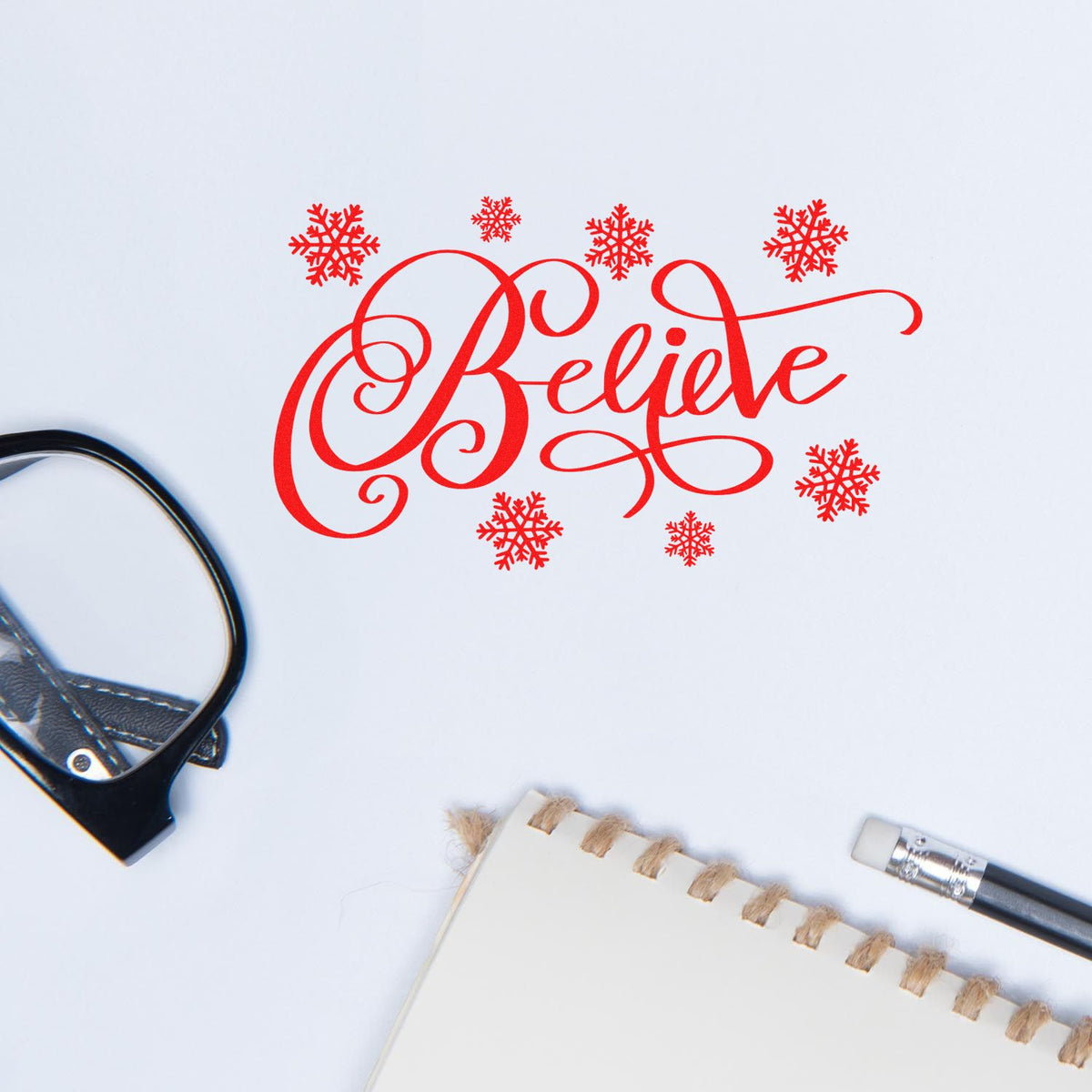 Red holiday stamp and seal reading Believe with snowflakes on white paper, beside notebook and tools.
