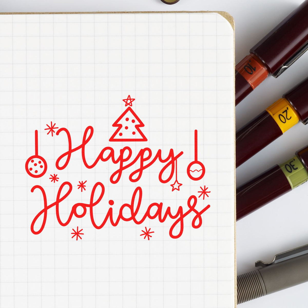 Red Happy Holidays stamp design with Christmas tree and ornaments on grid paper, beside stamp and seal pens with colored bands.