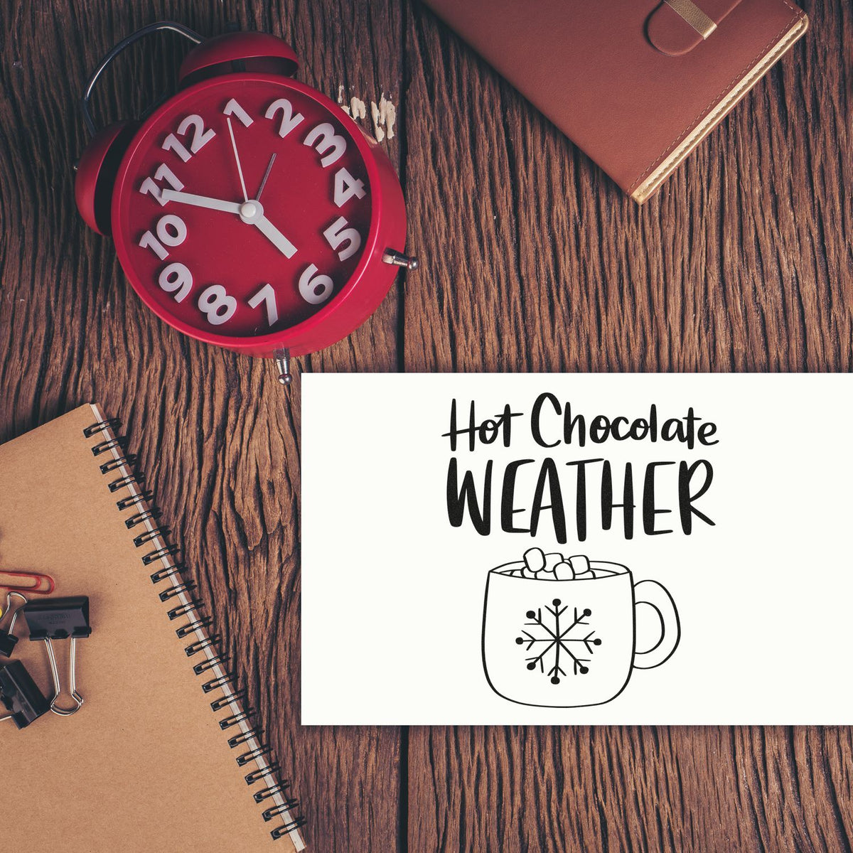 Red alarm clock with black-and-white Hot Chocolate Weather stamp and seal on white card, on rustic wood with tan notebook.