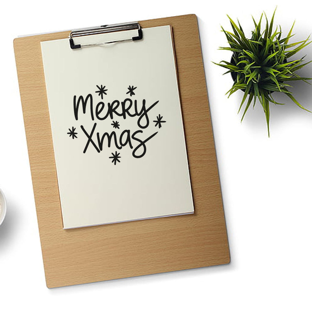 Black Merry Xmas stamp and seal design on white paper clipped to a wooden clipboard, with a green plant.