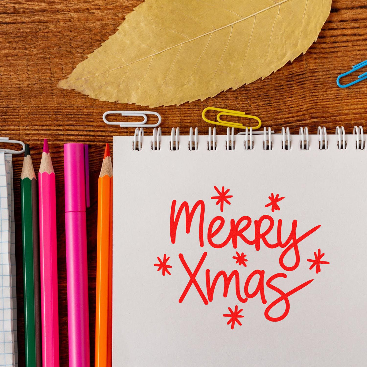 Red Merry Xmas stamp/seal design on spiral notebook, with colorful pencils, paper clips, and a gold leaf on wood.