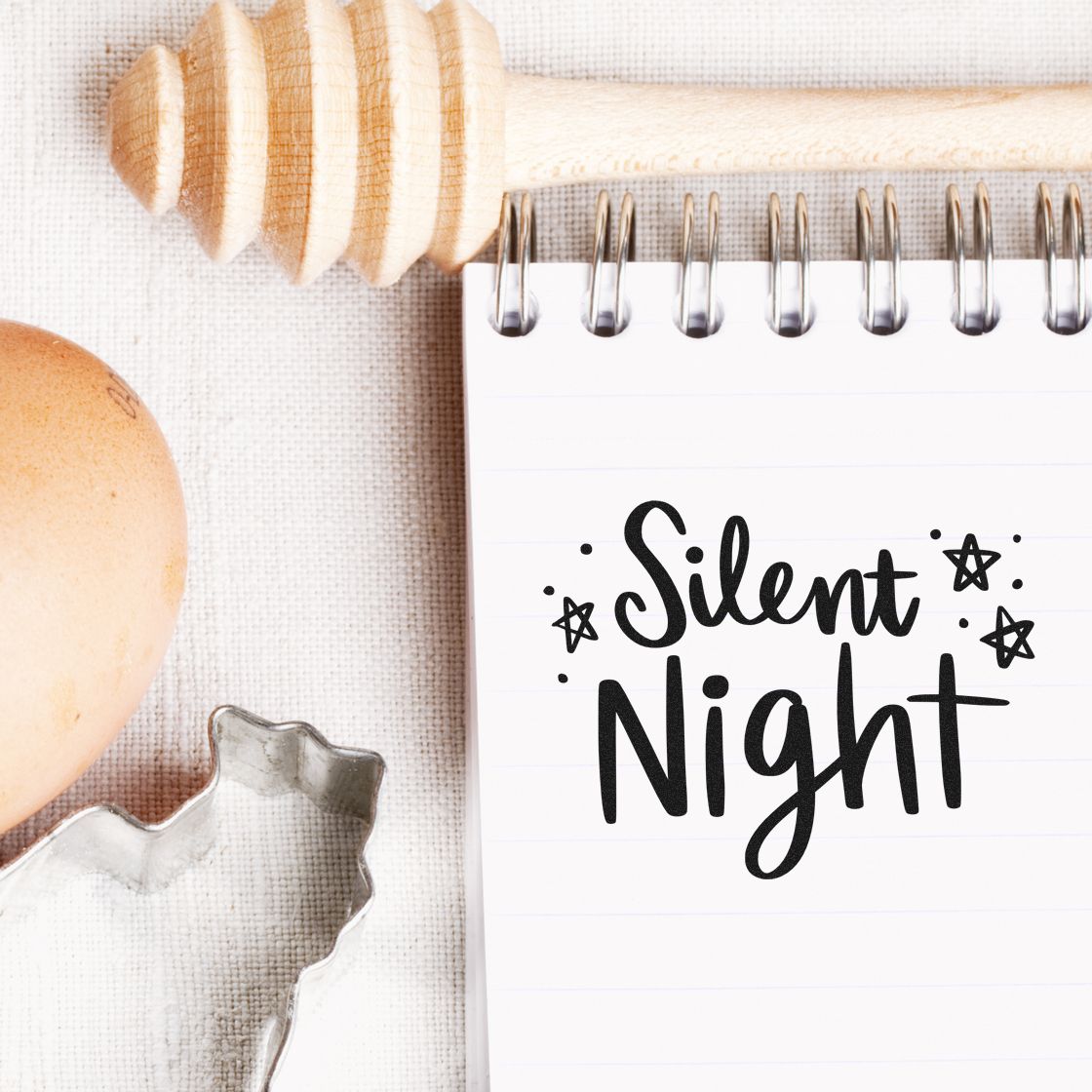 Silent Night stamp design on spiral notebook beside a wooden seal stamp, egg, and metal cutter on neutral fabric.