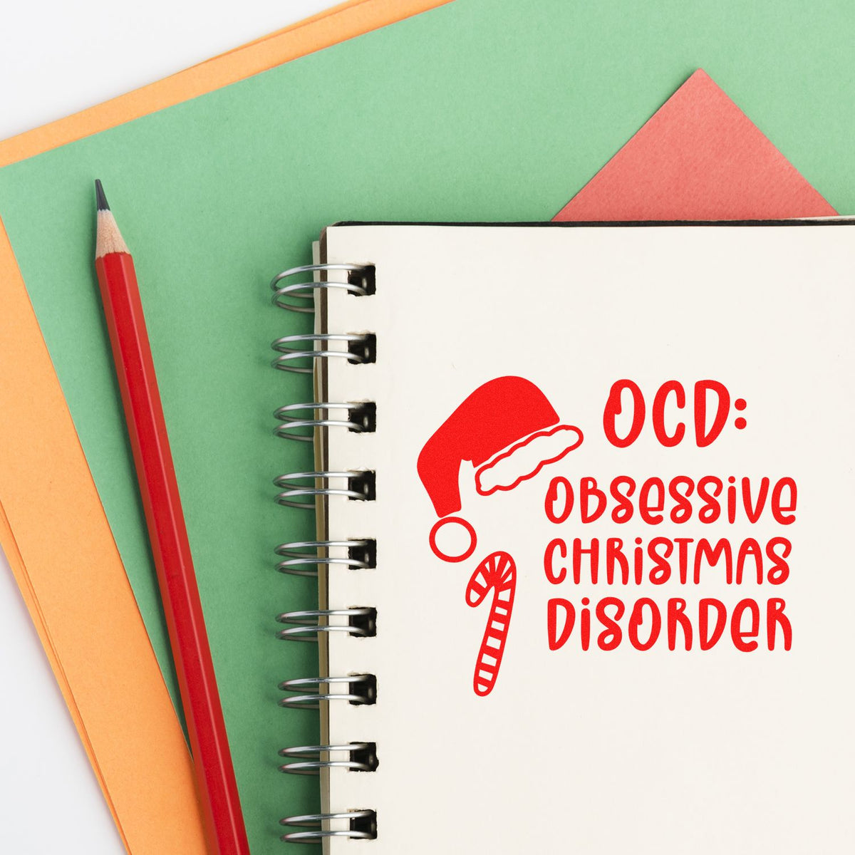 Red OCD: Obsessive Christmas Disorder stamp and candy cane seal on a spiral notebook, with a red pencil and green-orange paper