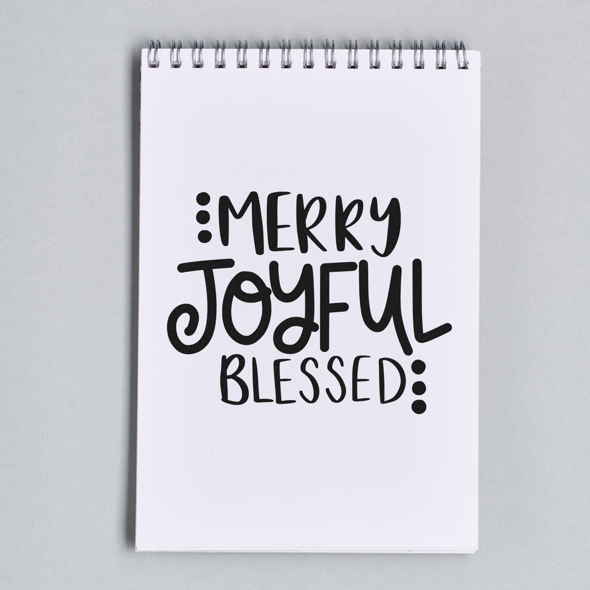Black Merry Joyful Blessed stamp and seal on a white spiral-bound notebook page with bold hand-lettered text.