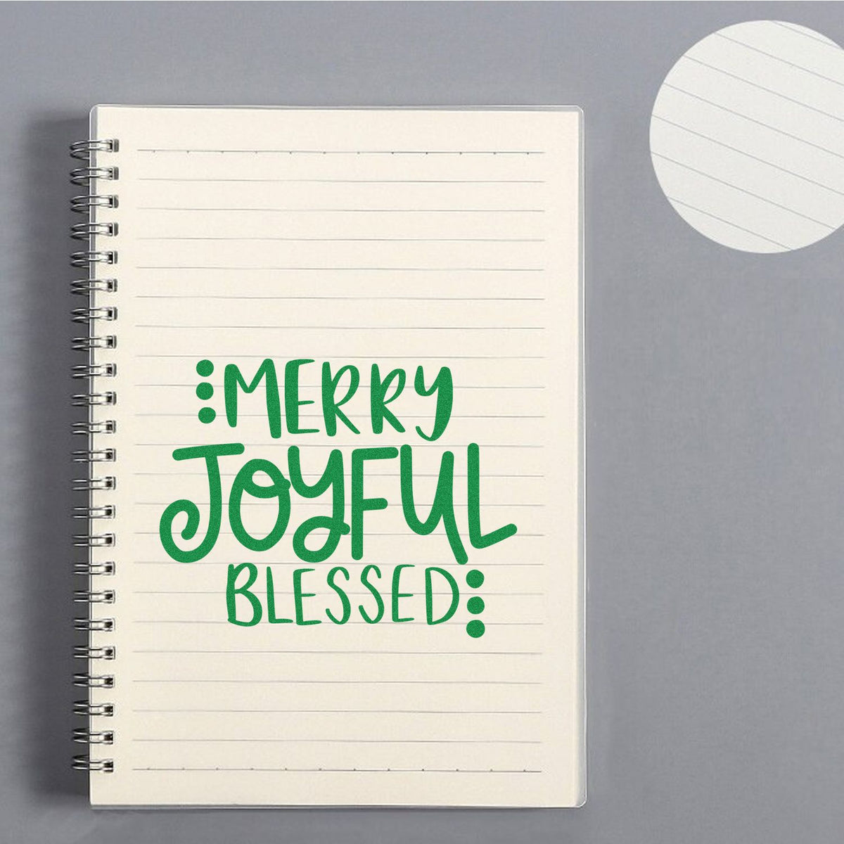 Green Merry Joyful Blessed stamp on cream spiral notebook with lined pages; white round seal at top right on gray background.