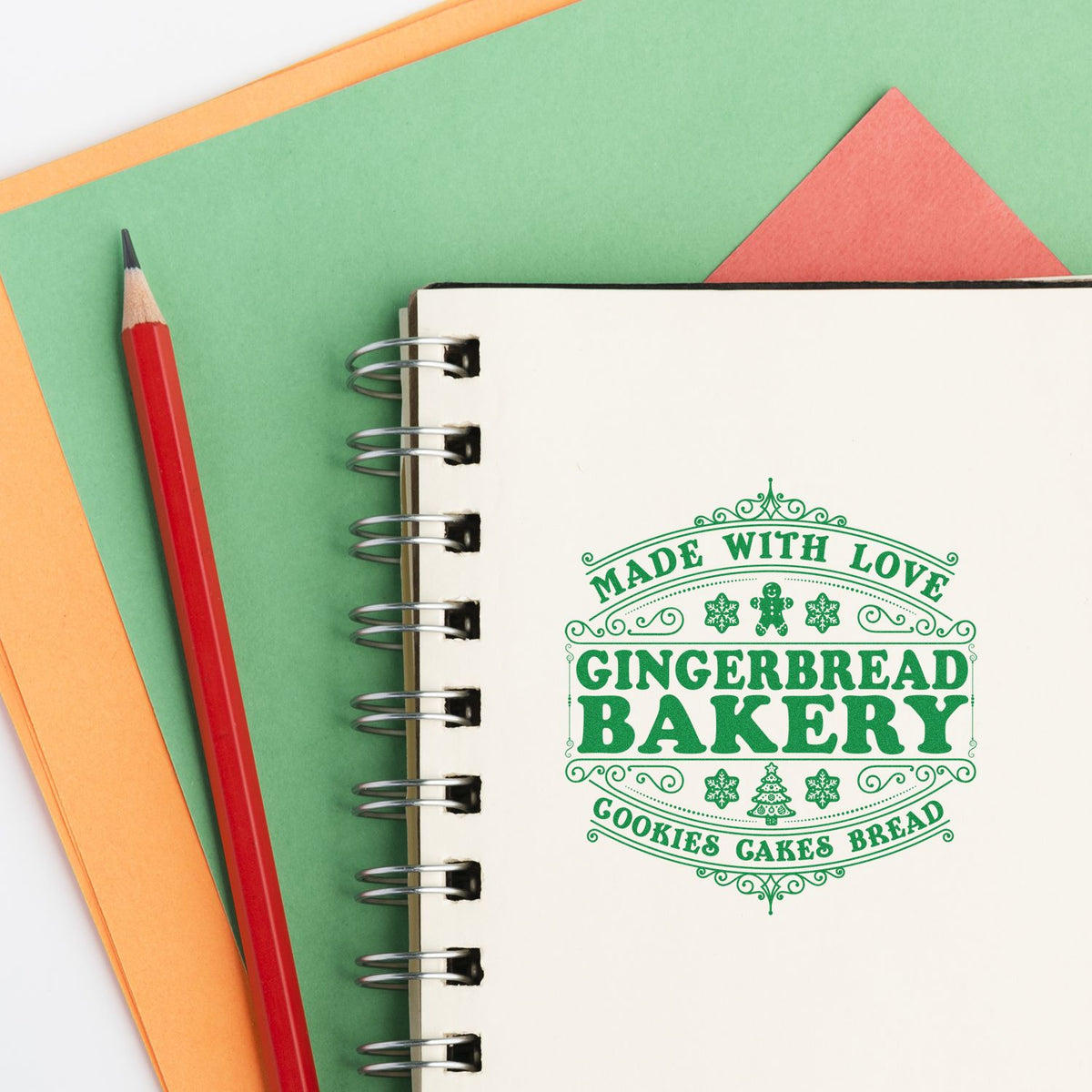 Green Gingerbread Bakery stamp seal on cream spiral notebook, with red pencil and pastel green, orange, and coral paper.
