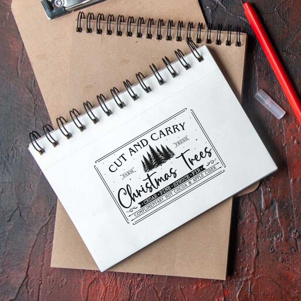 Custom holiday stamp and seal on white spiral notebook, featuring black Christmas tree design, kraft paper, red pencil, and dark wood.